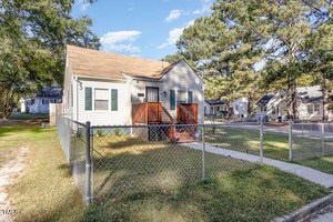 A small beige house with a tan roof sits behind a chain-link fence. It has a front porch with wooden steps and railings. Trees and a few other houses are visible in the background under a clear blue sky.