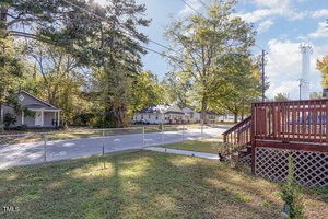 A suburban neighborhood featuring a view of a few houses and a street, surrounded by tall trees. A chain-link fence runs along the property with a wooden deck in the foreground. It's a sunny day with scattered clouds in the sky.