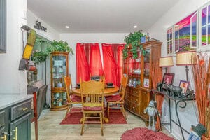 A cozy dining room with red curtains and a wooden table surrounded by chairs. The room is decorated with plants, framed pictures on the walls, and various ornaments, creating a warm and inviting atmosphere.