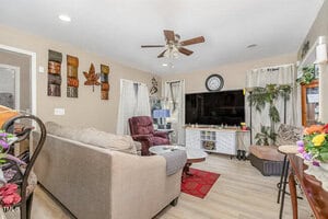 A cozy living room with a ceiling fan, beige walls, and light wood flooring. It features a large TV on a white stand, a brown sofa, and a purple recliner. Colorful wall art and potted plants add a touch of nature to the space.