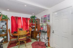 A cozy dining room with a wooden table and chairs. Red curtains cover the window, and various plants and framed pictures decorate the space. A cabinet with display items stands next to a white door. Bright lighting enhances the room's warmth.