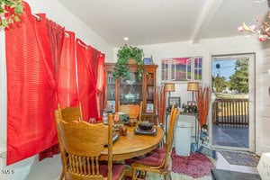 A cozy dining area with a wooden table and chairs, decorated with red curtains and a china cabinet filled with dishes. Plants and photos adorn the room, and a door opens to a porch with a view of trees and power lines outside.