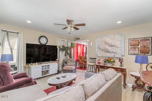 A cozy living room with a ceiling fan, a large TV, a sofa, and armchairs. The walls feature various artworks and a clock. A small dining area is visible through a doorway, with red curtains adding a pop of color.