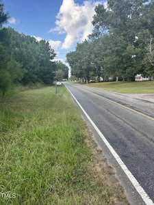 A rural road flanked by grass and trees on a sunny day. The road stretches into the distance with a clear sky and scattered clouds above. A sign is visible on the side of the road.