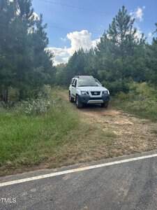A white SUV is parked on a dirt path surrounded by green trees under a partly cloudy sky. The vehicle is near a paved road, and the setting appears to be a rural, forested area.