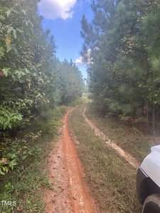 A narrow dirt path winds through a lush forest under a partly cloudy sky. The greenery of the trees lines the path, which shows signs of recent travel. A glimpse of a white vehicle is visible on the right side of the image.