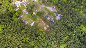 Aerial view of a forested area with a cluster of houses nearby. A clearing is marked with a red outline in the center of the image, surrounded by dense green trees. Roads and other buildings are visible in the upper part of the image.