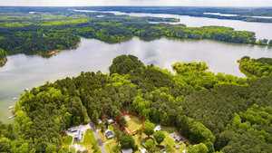 Aerial view of a lush, green landscape with a large lake surrounded by dense forests. A small residential area is visible in the foreground, with one plot outlined in red. The sky is overcast, casting a muted light over the scene.