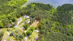 Aerial view of a lush, green forested area with a lake in the background. A suburban neighborhood is visible with several houses. A plot of land is outlined in red, bordered by trees, indicating a specific property.