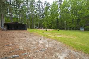 A rural backyard featuring a green lawn bordered by tall pine trees. A simple metal carport is on the left. In the distance, there is a playground with a swing set and a small shed on the right side. A dirt area is in the foreground.
