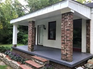 Located on 3111 Tungsten Mine Road in Bullock, this small house with a white exterior features a covered porch supported by brick columns. The porch boasts a dark gray painted floor. Surrounding the inviting entrance are landscaped areas with grass and plants, and a lantern-style light hangs by the white door.