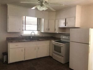 A kitchen with white cabinets, a ceiling fan, and a window above the sink. It includes a white refrigerator and a stove with an oven. The countertop is dark and the floor is tiled.