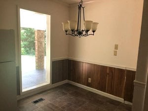 A small dining area with tiled flooring, a hanging light fixture, and wood-paneled walls. A large vertical window provides natural light, showing a view of a covered patio and greenery outside.