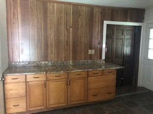 A room featuring a wooden paneled wall and a granite countertop. Below the countertop, there are several wooden cabinets and drawers. A door is visible to the right, next to a small window. The floor is tiled in a dark color.