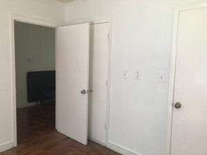 A minimal interior with white walls features three closed doors. Two doors are side by side, and a third door on the left is partially open, revealing a glimpse of a dark room and a couch. The floor is wooden.