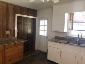A kitchen with wooden and white cabinets, granite countertops, and a tiled floor. There is a window with blinds above the sink and a ceiling fan above. A door with a glass window is on one wall, and wood paneling is on another wall.