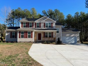 A charming two-story house at 41 Sandpipers Lane in Manson boasts beige siding and red shutters under a clear blue sky. It features a welcoming front porch, a red door, and an attached two-car garage, all surrounded by trees and a well-maintained lawn and driveway.