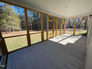 A spacious screened-in porch with a concrete floor and wooden frame overlooks a grassy yard and dense trees. Sunlight streams through, casting shadows on the floor.