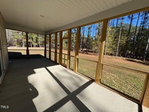 A view of a screened-in porch with a concrete floor, wood framing, and large windows. The porch overlooks a grassy yard with trees in the background, under a clear blue sky.