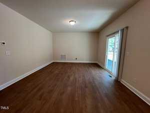 An empty room with wooden flooring and beige walls. A ceiling light fixture is centered above. Sliding glass doors lead to an outdoor area, allowing natural light to enter.