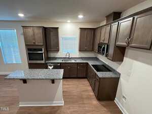 Modern kitchen with dark wood cabinets, granite countertops, and an island. Features include a built-in oven, microwave, stovetop, and stainless steel sink. Light hardwood floor and window with blinds add brightness.