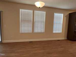 A room with three tall windows covered by closed blinds, beige walls, and wooden flooring. A ceiling light fixture is centered above the empty space. The room appears well-lit with a modern and minimalist design.