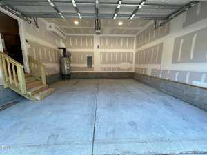 A clean, newly constructed garage with unpainted drywall and a concrete floor. Wooden stairs lead to a door on the left. A water heater is installed at the back corner. The garage door is open, letting in natural light.