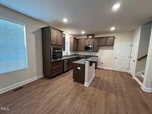 A modern kitchen with wooden cabinets, dark countertops, and stainless steel appliances. It features an island with additional workspace and storage. The room has hardwood floors, recessed lighting, and a window with blinds.