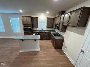 A modern kitchen with brown cabinets and granite countertops. It features an island with a sink, built-in oven, microwave, and ample storage. The room is well-lit with windows covered by blinds and has wooden flooring.