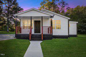 Located at 406 Harriett Street in Henderson, this charming small white house boasts a black door and red trim. A welcoming front porch and steps lead to a sidewalk, framed by lush green grass and trees under a vibrant purple and pink sunset sky.