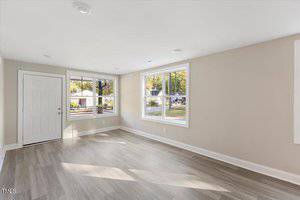 A bright, empty room with light beige walls and large windows letting in natural light. The floor is covered in light wood laminate. A white door leads to an outdoor area with trees and houses visible through the windows.