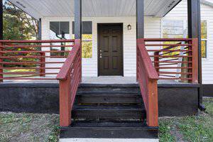 A front entrance of a house featuring a dark brown door, white siding, and black trim. The porch has wooden railings painted in red and black stairs leading up. Surrounding greenery is visible in the background.