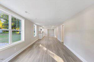 A bright, empty room with light wood flooring and beige walls. Large windows on the left bring in natural light. The room extends into a kitchen area with white cabinets. Doorways on the right lead to other rooms. Trees are visible outside.