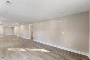 Spacious empty room with light wood flooring and neutral-colored walls, featuring recessed lighting and multiple doorways leading to other areas. A large window casts natural light onto the floor, enhancing the open, airy feel.