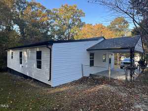 A white, single-story house with a metal roof and attached carport is surrounded by trees. The yard is fenced, and a car is parked in the driveway. Fallen leaves cover parts of the ground, indicating an autumn setting.