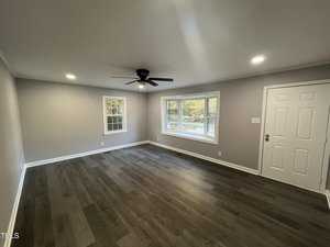 A spacious, empty room with gray walls and dark wood flooring. It features a white door, large windows, recessed lighting, and a black ceiling fan. Natural light enters through the windows, creating a bright and airy atmosphere.