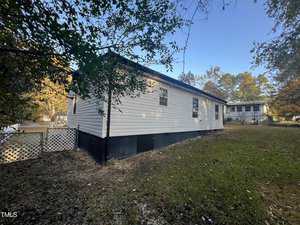 A single-story white mobile home with a dark foundation sits on a grassy lot. A white lattice fence borders one side. Trees with fall foliage are in the background under a clear blue sky.
