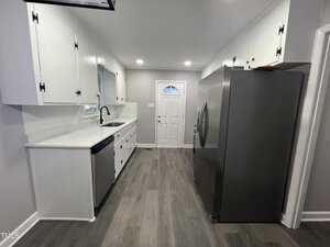 A modern kitchen with white cabinets, a stainless steel sink, and a large silver refrigerator. The floor is gray wood, and there's track lighting on the ceiling. A white door with a decorative window is at the end of the room.