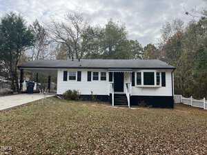 Nestled at 1910 Willow Lane, a small white house with black shutters graces a leaf-covered lawn. It features a carport on the left, steps leading to the front door, and trees standing tall in the background beneath Henderson's cloudy sky.