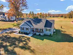 An aerial view showcases a modern, single-story house at 3217 Satterwhite Point Road in Henderson. The home features a gray exterior and dark roof, complemented by a front porch with chairs. It's nestled on open land with scattered trees under the clear blue sky.