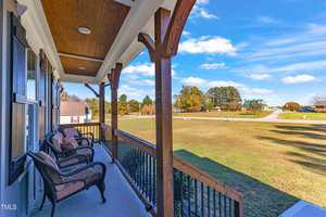 A porch with wicker chairs overlooks a spacious lawn. The view includes a clear blue sky, scattered clouds, and a few distant houses surrounded by trees. The porch features wooden beams and a railing.