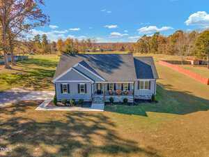 A charming single-story house with a large front lawn and a spacious driveway, surrounded by open fields and scattered trees under a clear blue sky with some clouds. The house has a welcoming front porch and is set in a rural landscape.