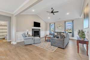 A cozy living room with light wood flooring, a gray sectional sofa, and an armchair facing a fireplace with a TV above. A large clock hangs near two windows with sheer curtains, and a small side table features a potted plant.