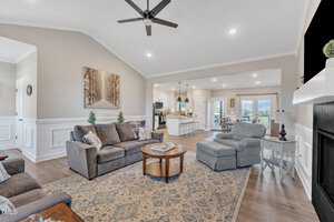 Modern living room with neutral tones, featuring a gray sofa and armchair, a round wooden coffee table on a patterned rug, and a fireplace. The open layout leads to a kitchen with a large island and a dining area with a clear view of the outdoors.