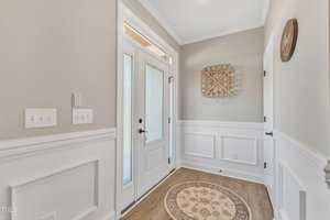 A bright entryway with a glass-paneled door, white wainscoting, and beige walls. A round rug with a floral pattern is on the floor. A decorative wall piece and a clock are mounted on the walls.