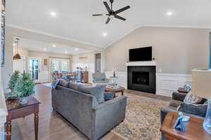 A spacious living room with light-colored walls and wood flooring. It features a gray sofa, armchairs, a fireplace with a flat-screen TV above, and decorative plants. A connected dining area is visible in the background. A ceiling fan hangs overhead.