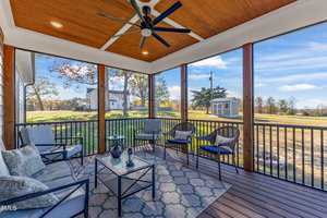 A screened porch with patio furniture including chairs and a table with a teapot and cups. The porch has a wooden ceiling with a fan. The view features a yard, trees, and a distant white house under a blue sky with scattered clouds.