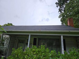 A small, white house with a dark roof, partially obscured by green shrubs. The front door is visible on the left, and a large window with drawn curtains is on the right. A brick chimney is on the roof's edge, surrounded by trees.