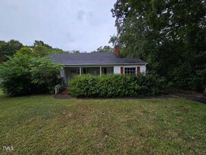 Nestled at 1530 Oakdale Circle in Henderson, this charming single-story house features a dark roof and white siding. A cozy front porch is partially concealed by dense shrubs, surrounded by lush green trees and a grassy lawn under a cloudy sky.