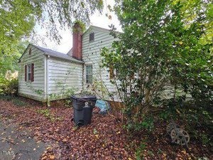 A weathered, white wooden house with red shutters partially hidden by overgrown bushes and trees. A wheelbarrow and a black trash bin are in front of the house, amidst fallen leaves on the ground. A brick chimney is visible on the roof.